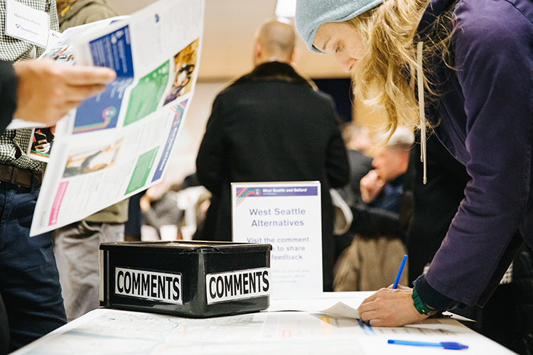 A person writes a comment on a sheet of paper adjacent to a comment collection box next to informational materials about West Seattle alternatives. 